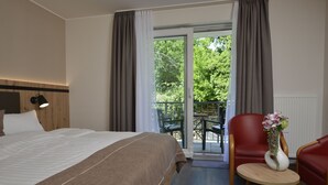 Camera d'albergo con un letto, un tavolino con un vaso, una sedia rossa e un balcone con vista sugli alberi.