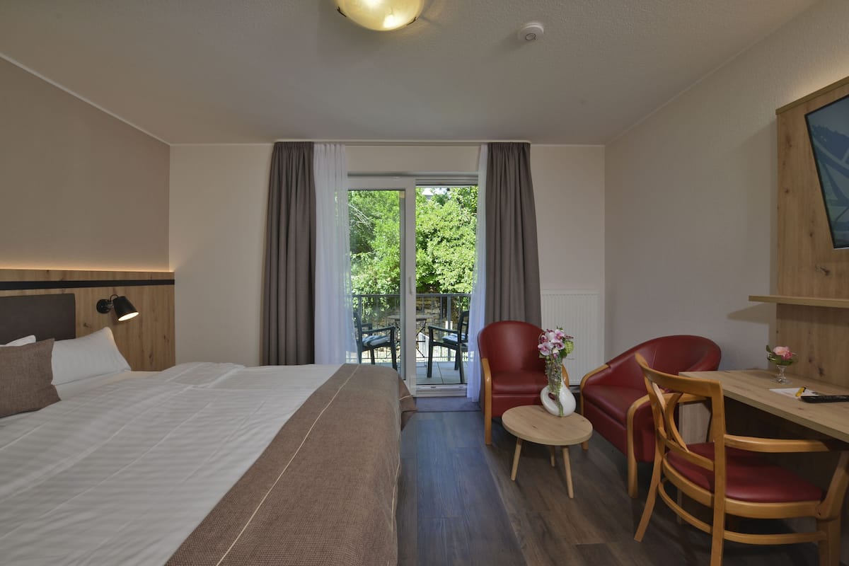 A hotel room with a bed, a desk, two chairs, a small table, and a view of the outdoors through a glass door.
