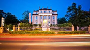 Un edificio grande e histórico con fachada simétrica, entrada central y un jardín formal con cerca.