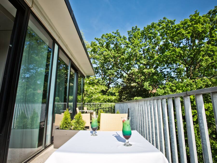 Een balkon met een tafel voor twee personen, twee drankjes en uitzicht op bomen.