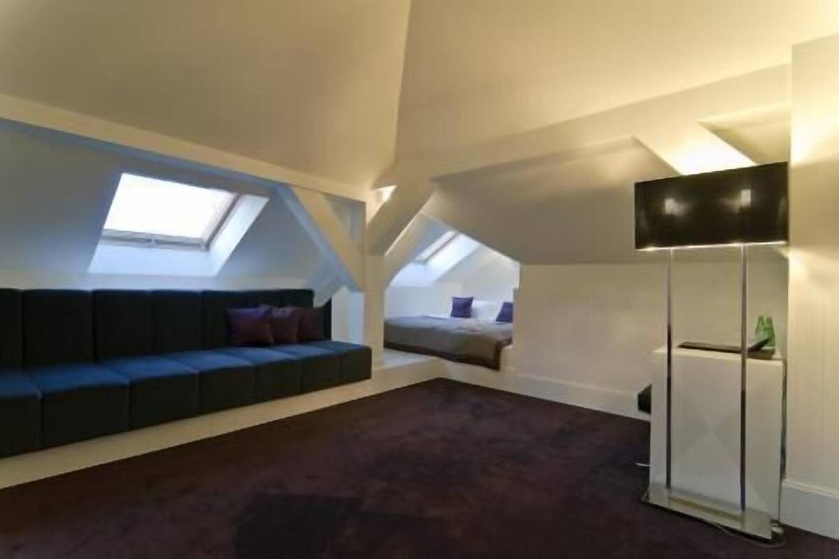 Ein Schlafzimmer im Loft-Stil mit hohen Decken, Dachfenstern, einem dunkelgrauen Sofa, einem Bett und einer Stehlampe.