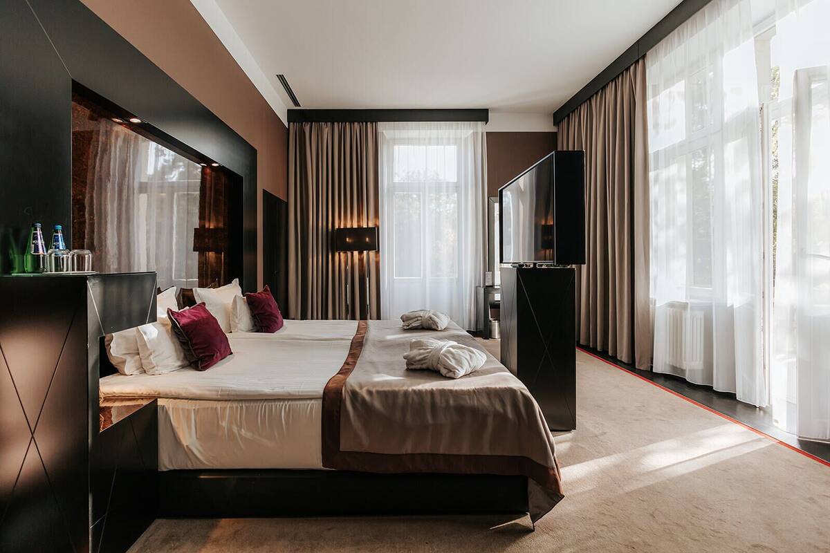 Ein modernes Hotelzimmer mit zwei Betten, einem Flachbildfernseher, einer Minibar und großen Fenstern mit transparenten Vorhängen.