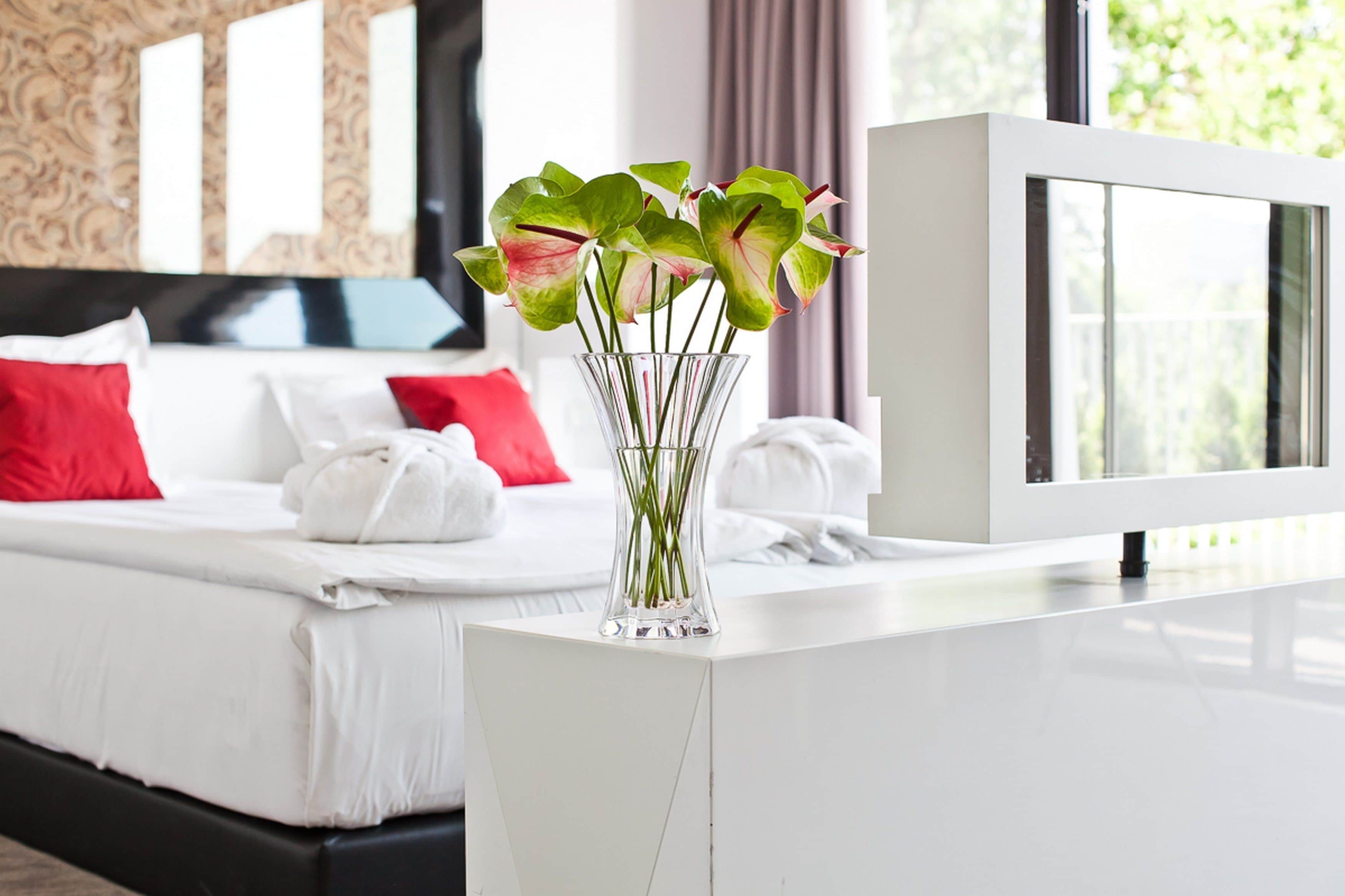 A modern hotel room with a bed, white linens, red pillows, a glass vase with flowers, and a large window.