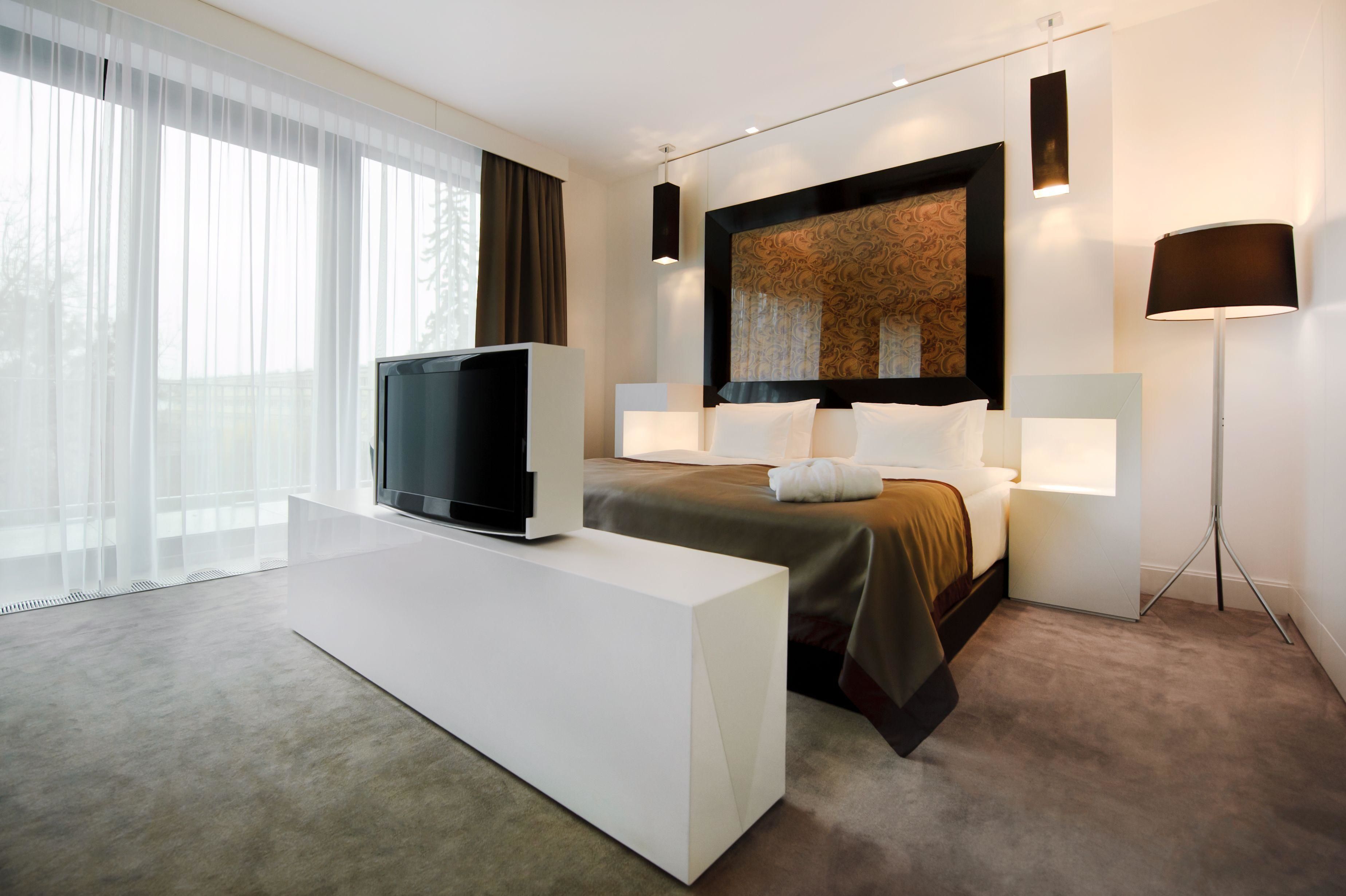 A modern hotel room with a large bed, a flat-screen TV, a white console table, and a large window with sheer curtains.