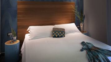 A neatly made bed with white linens, a decorative pillow, and a blue throw blanket. There are two bedside tables with decorative items and a vase of plants.