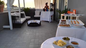 A rooftop terrace with a dining setup, including a table with food and drinks, seating arrangements, and a view of the cityscape.
