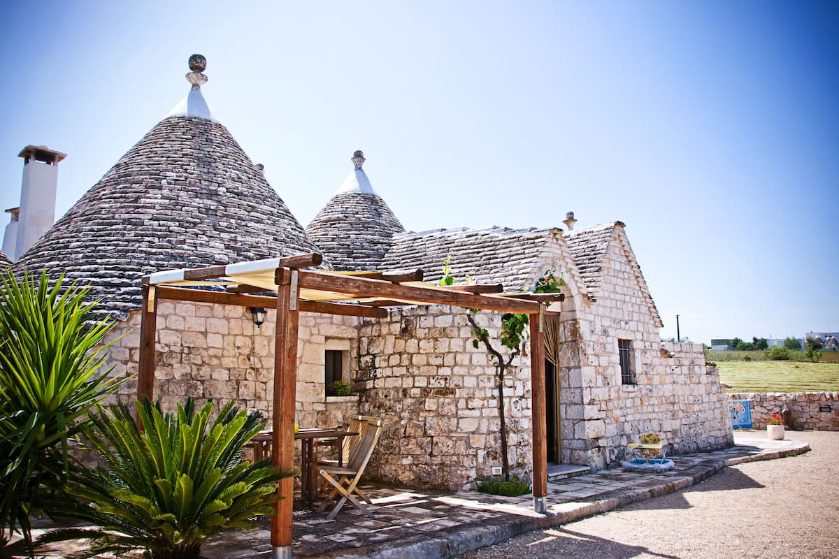 A stone building with conical roofs, a wooden pergola, and a small table and chair set outside.