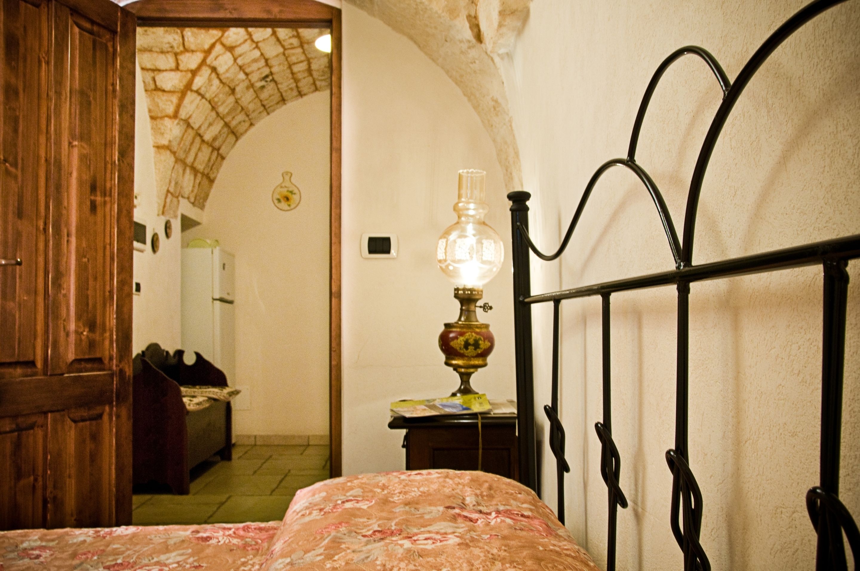 A bedroom with a bed, a wooden wardrobe, a lamp, and an arched doorway leading to another room.