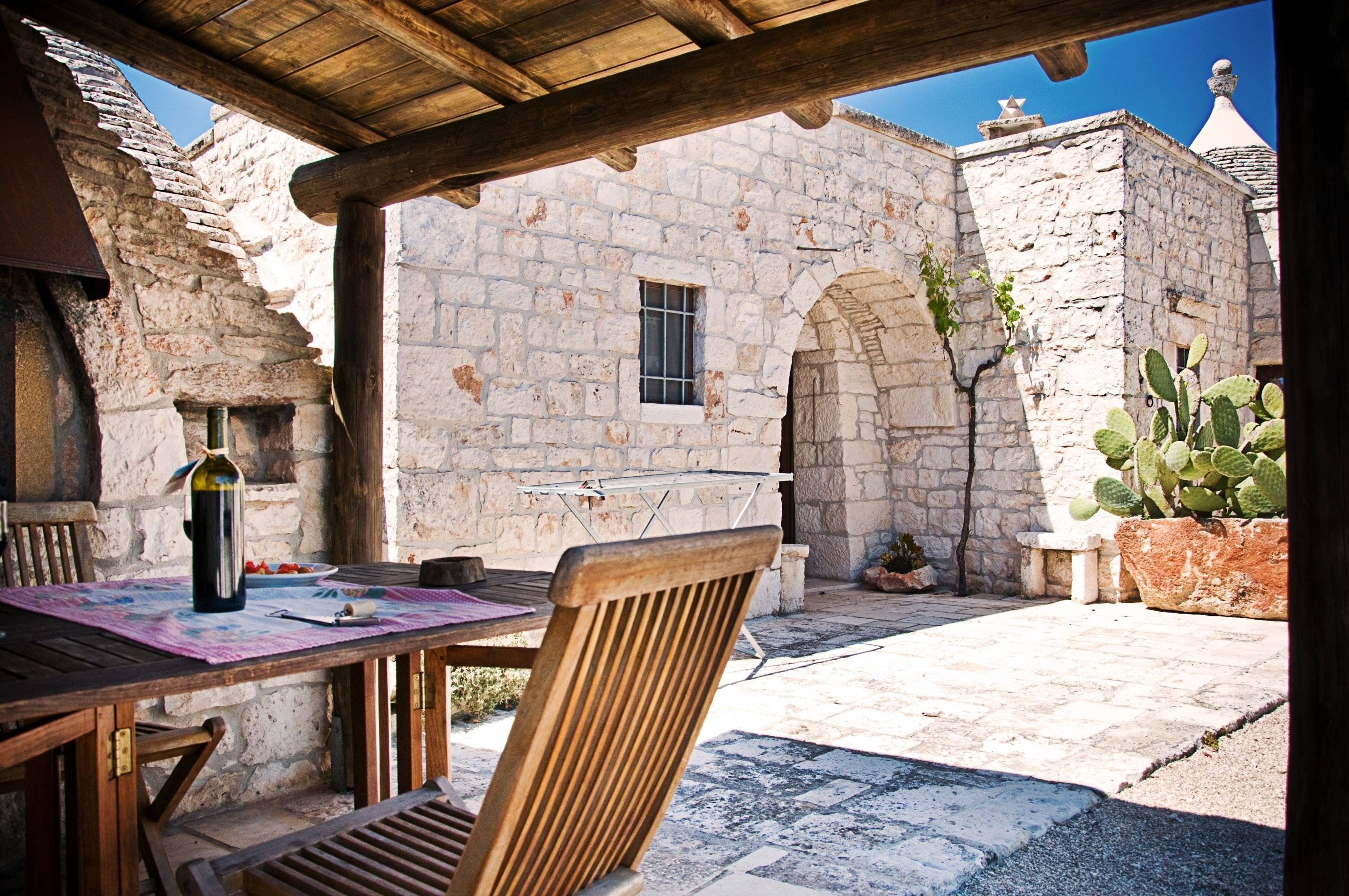 Un bâtiment en pierre avec une table et des chaises en bois, un cactus et une bouteille.