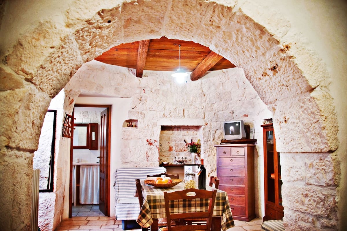 A rustic dining area with a stone archway, wooden ceiling beams, a table set with a checkered tablecloth, and a television on a cabinet.