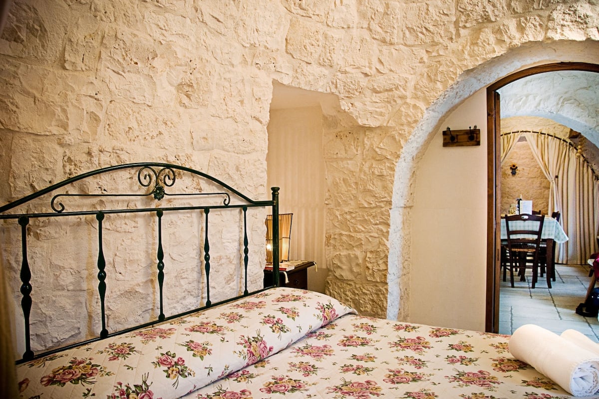 A bedroom with a bed, a stone wall, and an arched doorway leading to a dining area.