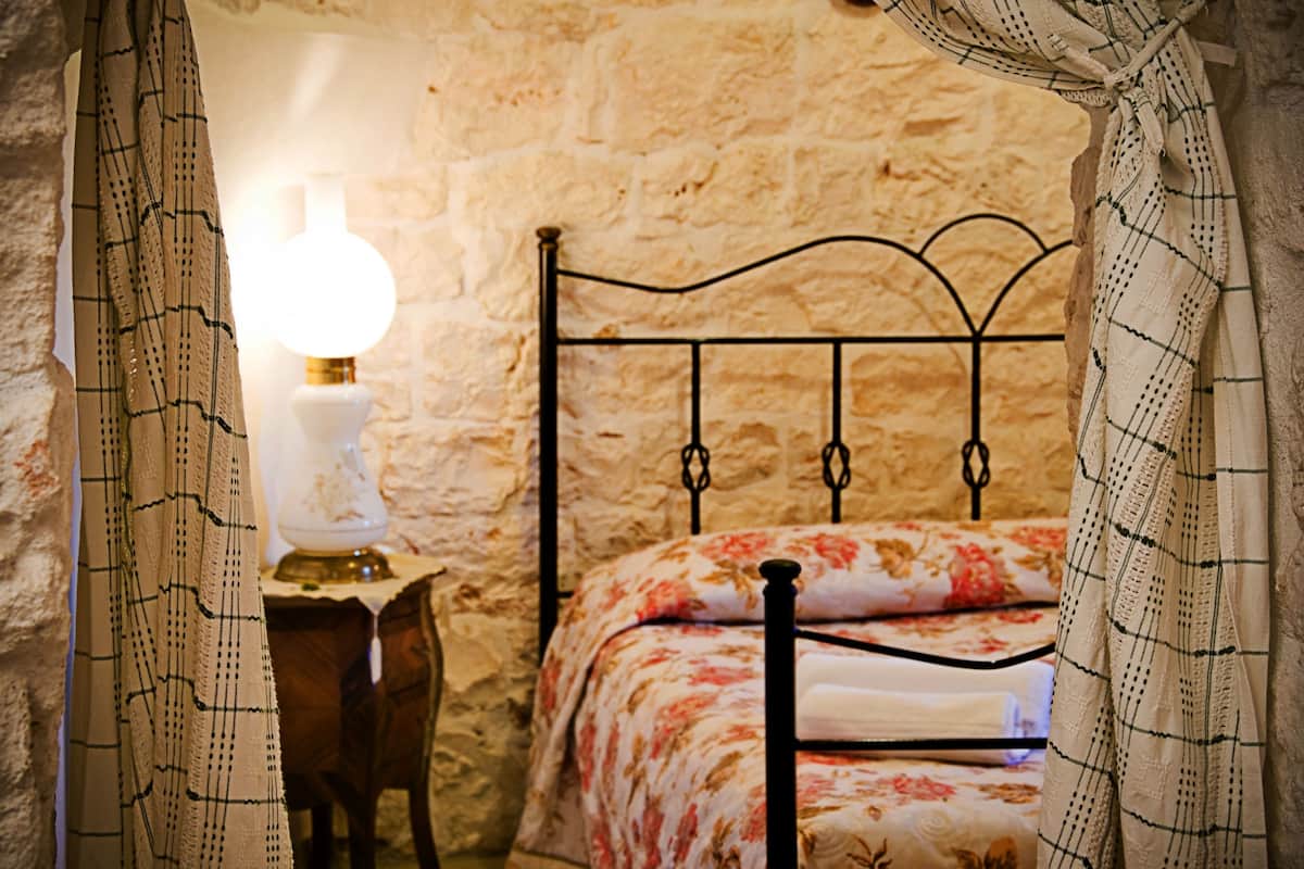 A room with a stone wall, a metal bed frame, a bedside table with a lamp, and a floral bedspread.