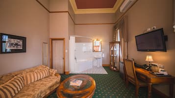 Une chambre d’hôtel comprenant un canapé, une table basse ronde, un bureau avec une lampe, une télévision, une salle de bain avec une baignoire et un tableau encadré au mur.