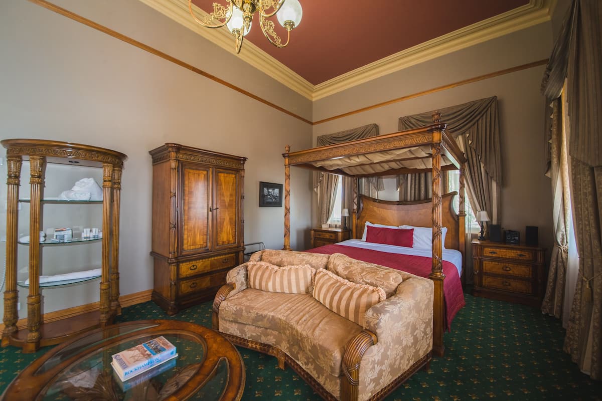 A bedroom with a four-poster bed, a wooden wardrobe, a glass-fronted cabinet, and a green carpeted floor.