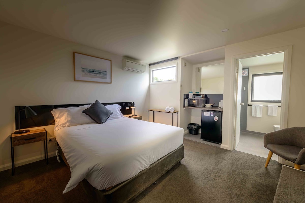 A hotel room with a bed, bedside tables, a desk, a chair, a kitchenette, and a bathroom.