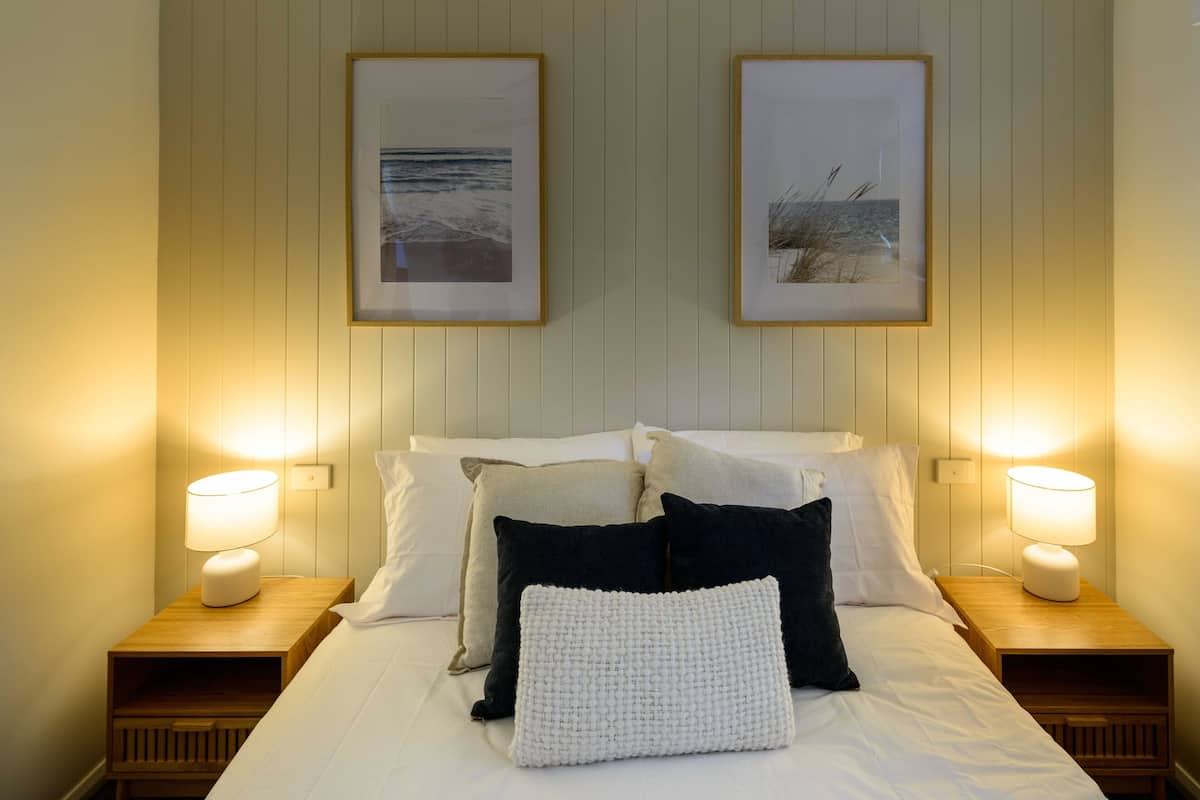 A neatly made bed with white and dark pillows, two framed pictures on the wall, and bedside lamps on wooden nightstands.