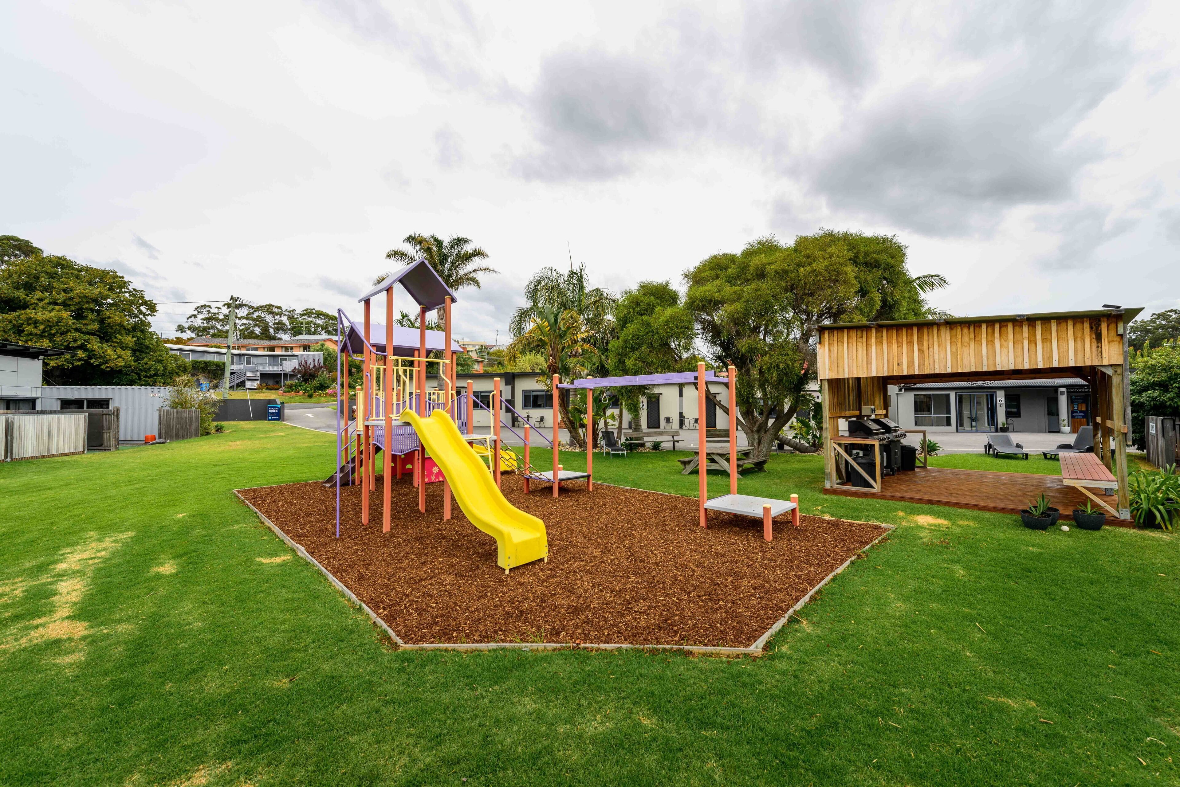 A playground with a yellow slide and a climbing structure in a grassy area with a wooden deck and outdoor seating.