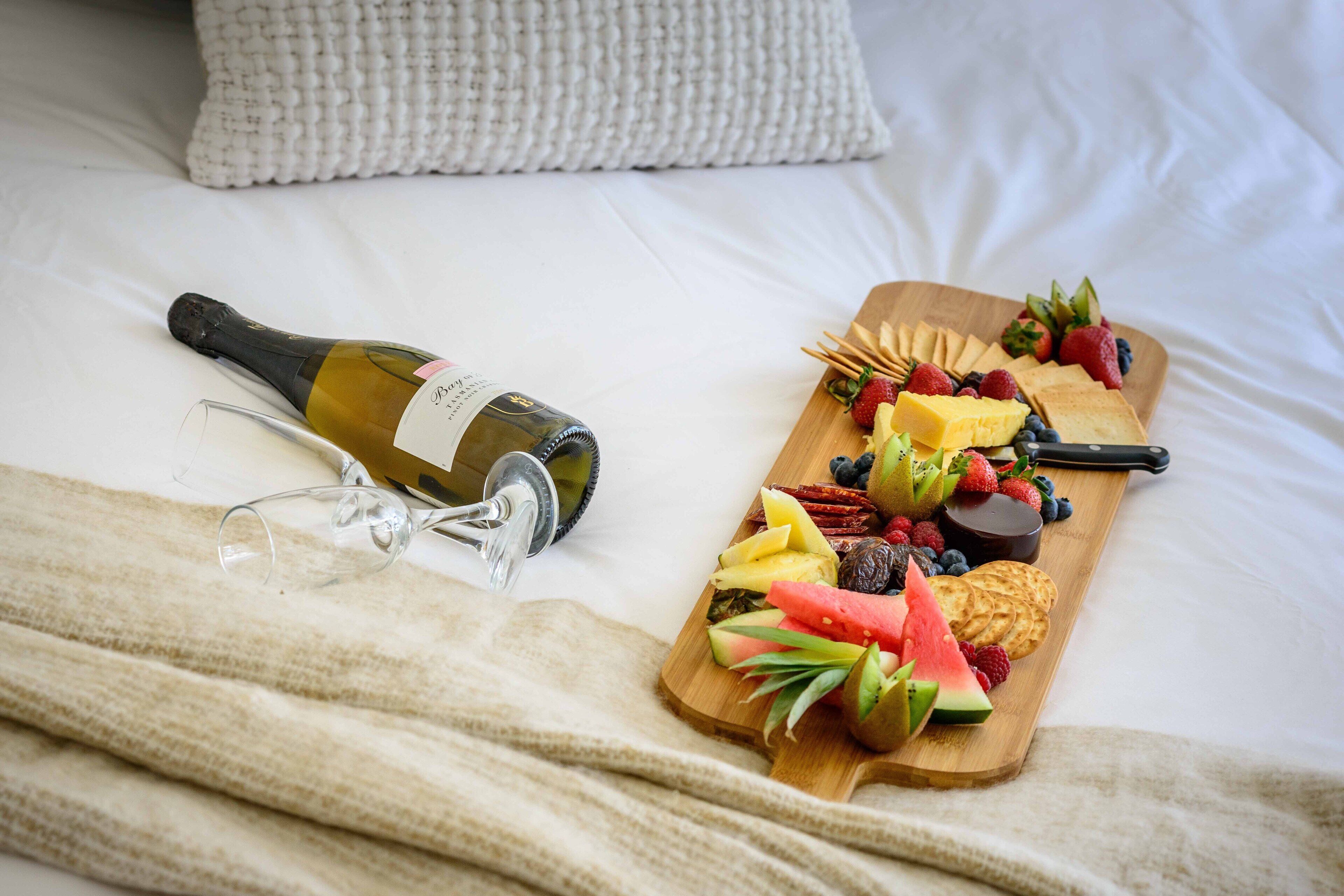 Una cama con una manta blanca, una botella de vino y una tabla de madera con frutas y aperitivos.