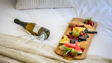 Una cama con una manta blanca, una botella de vino y una tabla de madera con frutas y aperitivos.