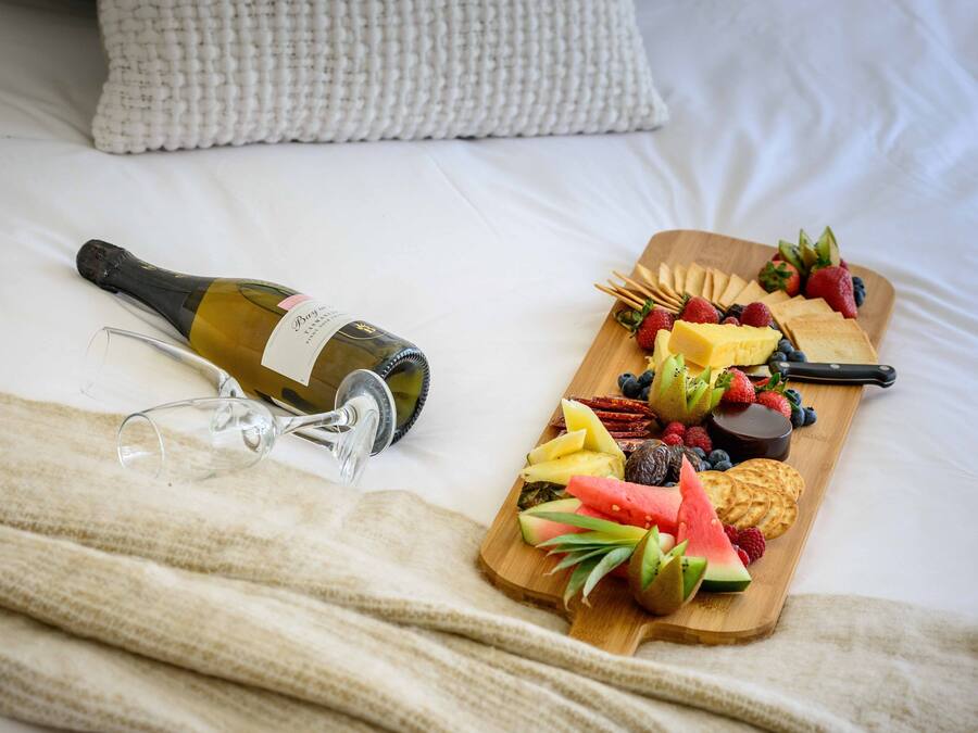 Una cama con una manta blanca, una botella de vino y una tabla de madera con frutas y aperitivos.
