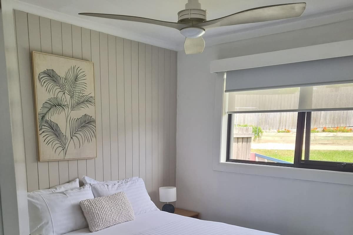 A bedroom with a bed, a ceiling fan, a window with a view of the outdoors, and a wall-mounted artwork.