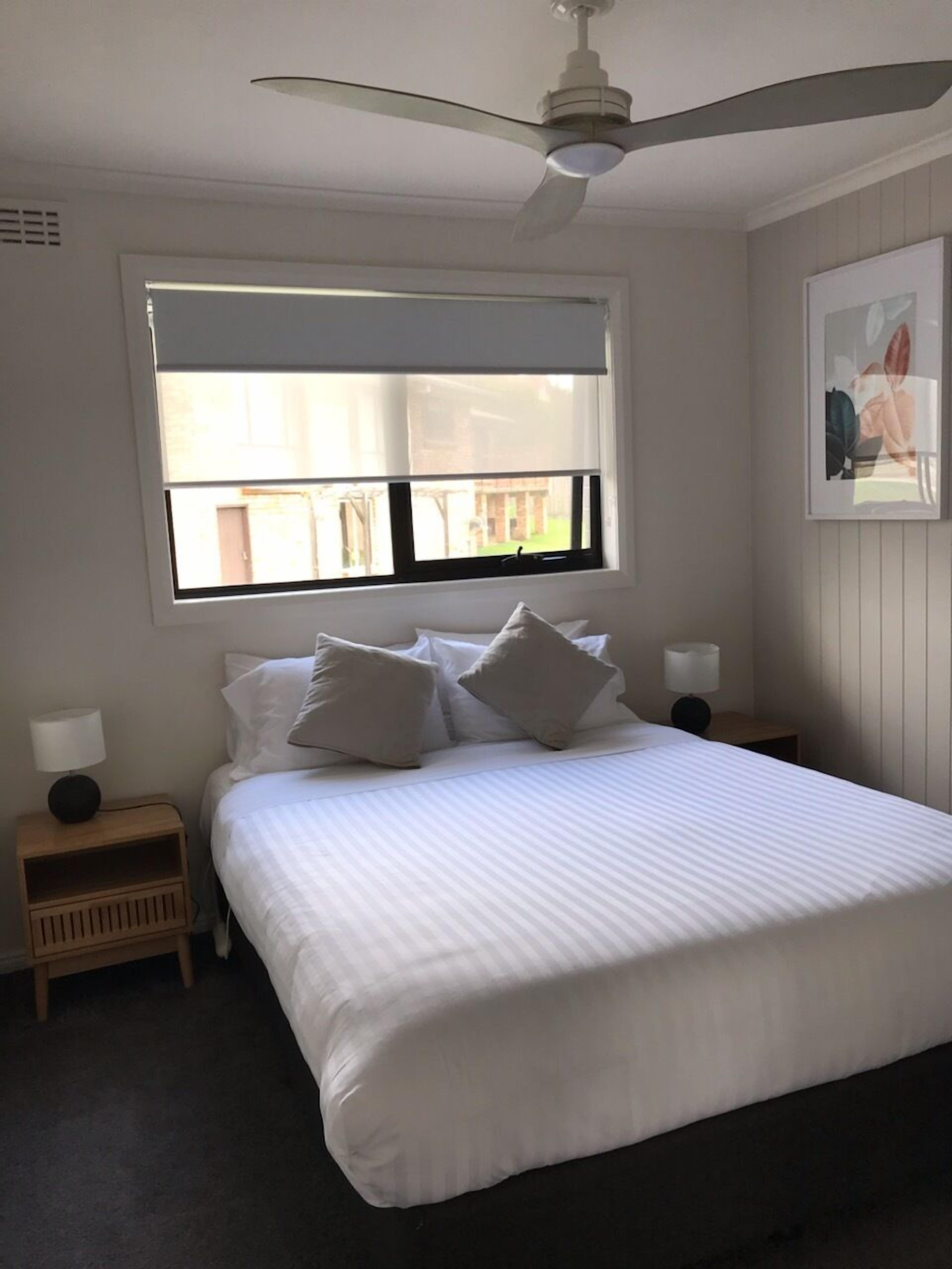 A neatly made bed with white linens and pillows, a ceiling fan, a window with blinds, and a small bedside table with a lamp.