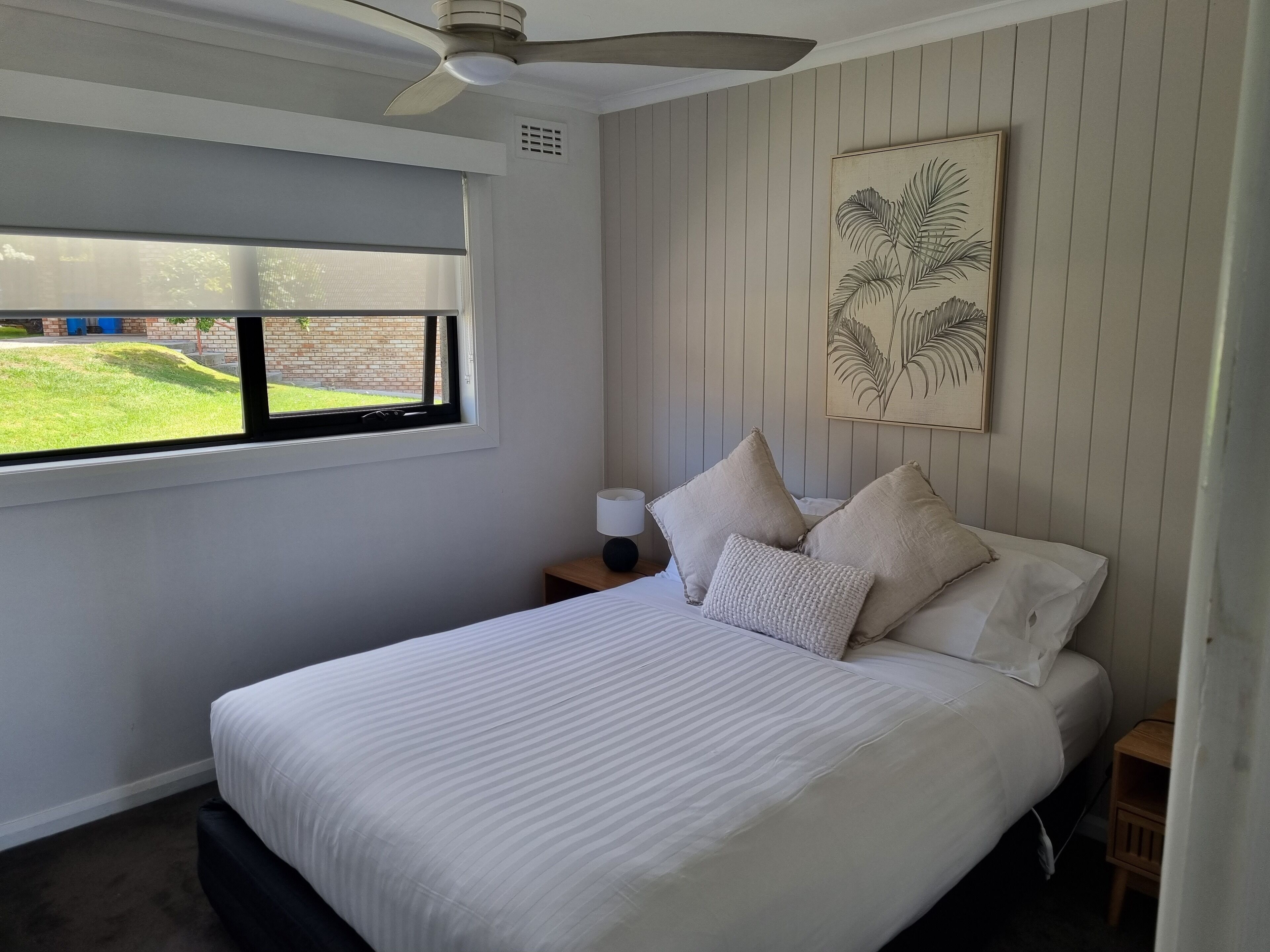 A bedroom with a large bed, a ceiling fan, a window with blinds, a nightstand with a lamp, and a wall-mounted artwork.