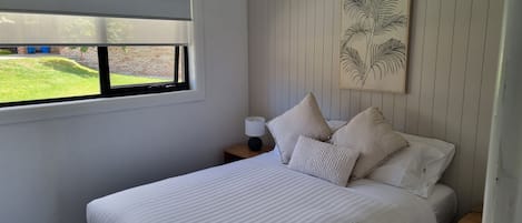 A bedroom with a large bed, a ceiling fan, a window with blinds, a nightstand with a lamp, and a wall-mounted artwork.