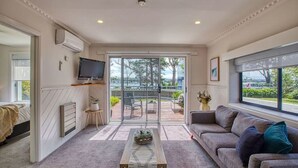 Una sala de estar moderna con sofá, una mesa de centro y televisión. Hay un ventanal corredera que da a un patio exterior con vistas a árboles y una carretera.
