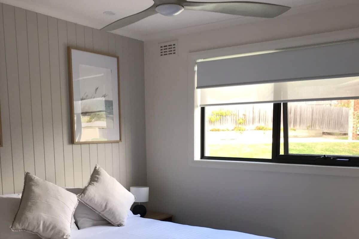A bedroom with a bed, a ceiling fan, a window with a view of the outdoors, and a framed picture on the wall.