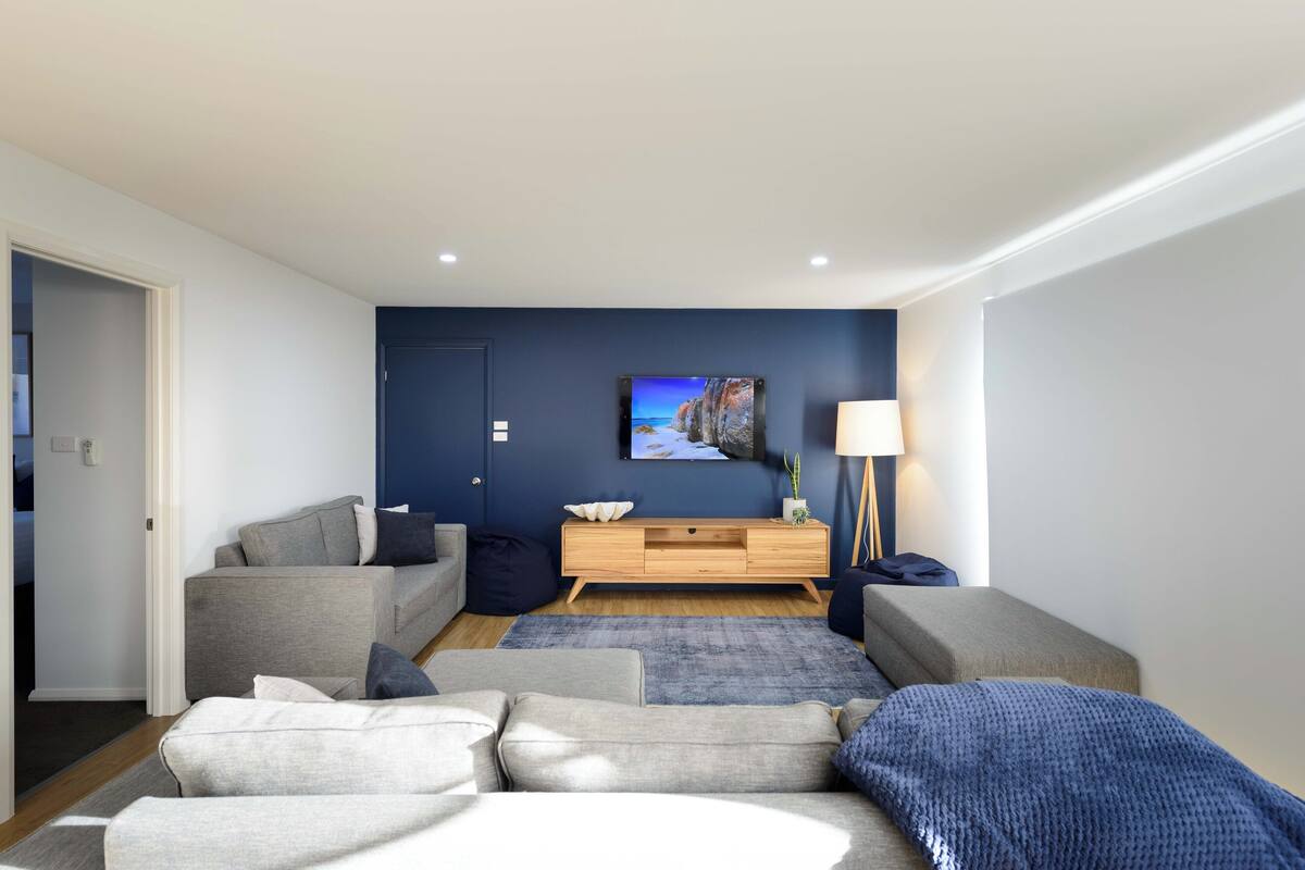 A modern living room with a grey sofa, a blue accent wall, a wooden TV stand, and a lamp.