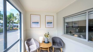 Una habitación con dos sillas grises, una mesita redonda y un jarrón con flores. Hay dos cuadros en la pared y un ventanal con vistas a palmeras y un edificio.