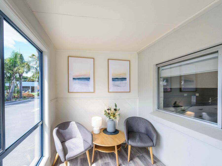 A room with two grey chairs, a small round table, and a vase with flowers. There are two framed pictures on the wall, and a large window with a view of palm trees and a building.