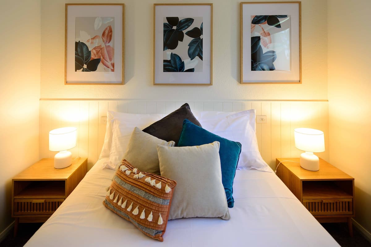 A neatly made bed with multiple pillows and a decorative throw, flanked by two bedside tables with lamps, in a room with framed artwork on the wall.