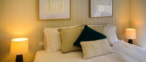 A neatly made bed with white linens, two pillows, and a throw blanket. There are two bedside lamps on wooden nightstands. Two framed artworks are hung on the wall above the bed.