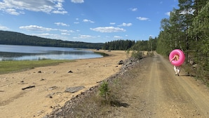Un chemin de terre mène à un lac, bordé d’une forêt d’un côté et surplombé d’un ciel bleu clair.