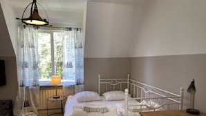 Une chambre à coucher avec un lit, une table en bois, une lampe, une fenêtre avec des rideaux et un radiateur.