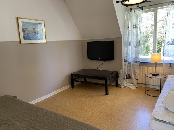 A room with a television mounted on the wall, a small table, a window with curtains, and a bed. - Garpgården (Garpenberg)