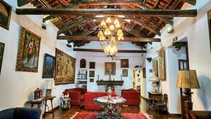 Ein traditionell eingerichteter Raum mit hohen Holzbalken, einem Kronleuchter, roten Sofas, einem gemusterten Teppich und verschiedenen gerahmten Kunstwerken.