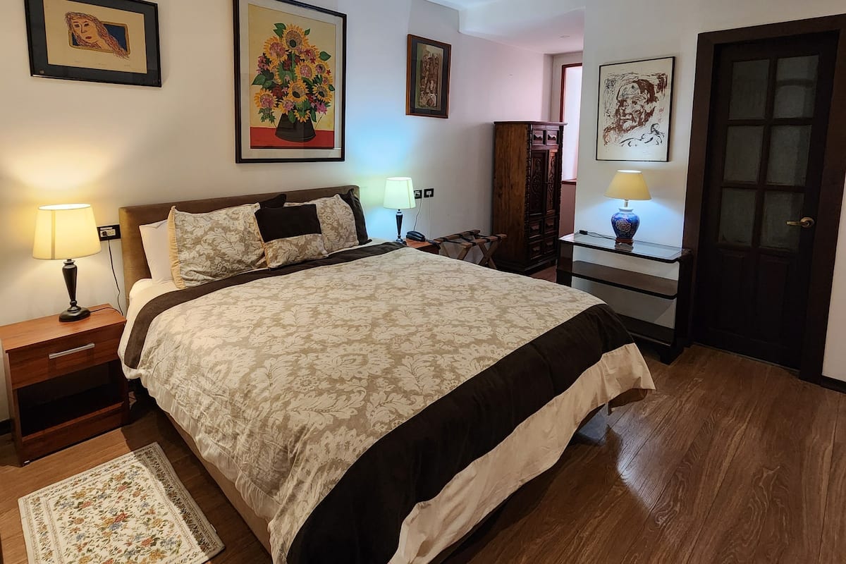A bedroom with a large bed, a nightstand, a lamp, a wooden dresser, and a door.