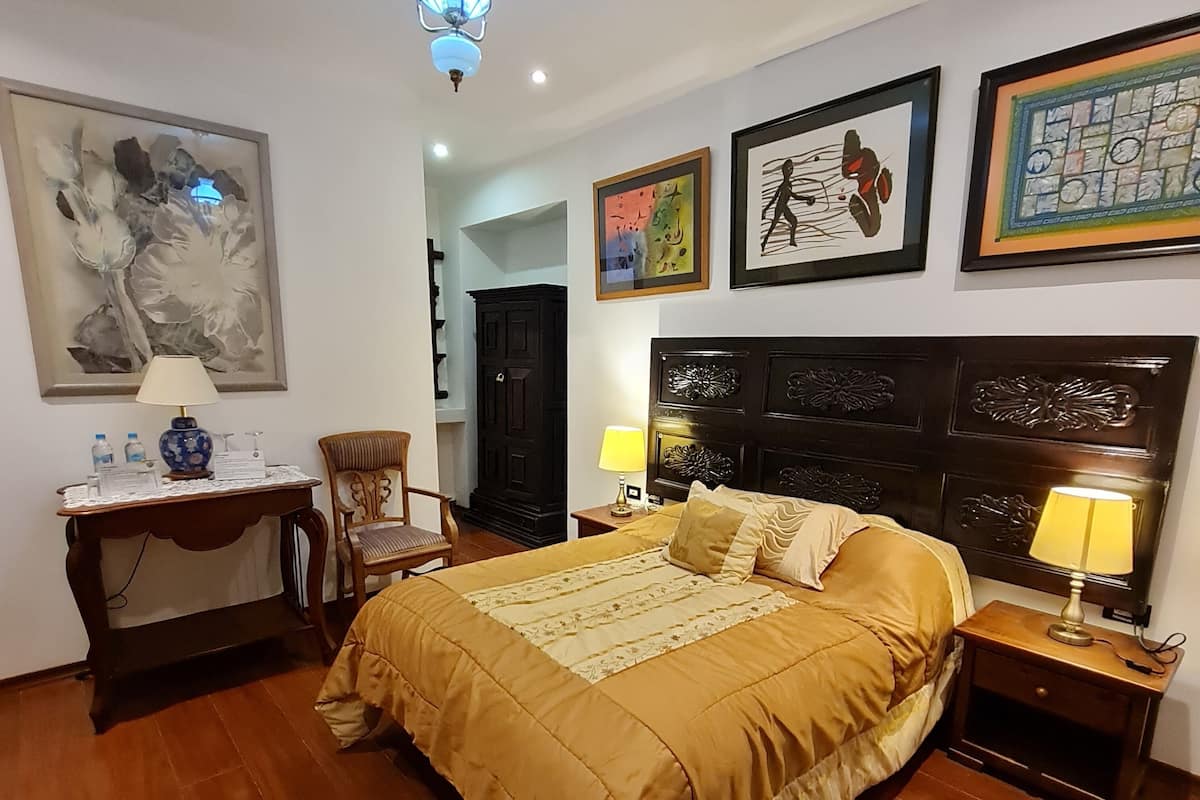 A bedroom with a large bed, a desk with a lamp, a chair, and artwork on the walls.