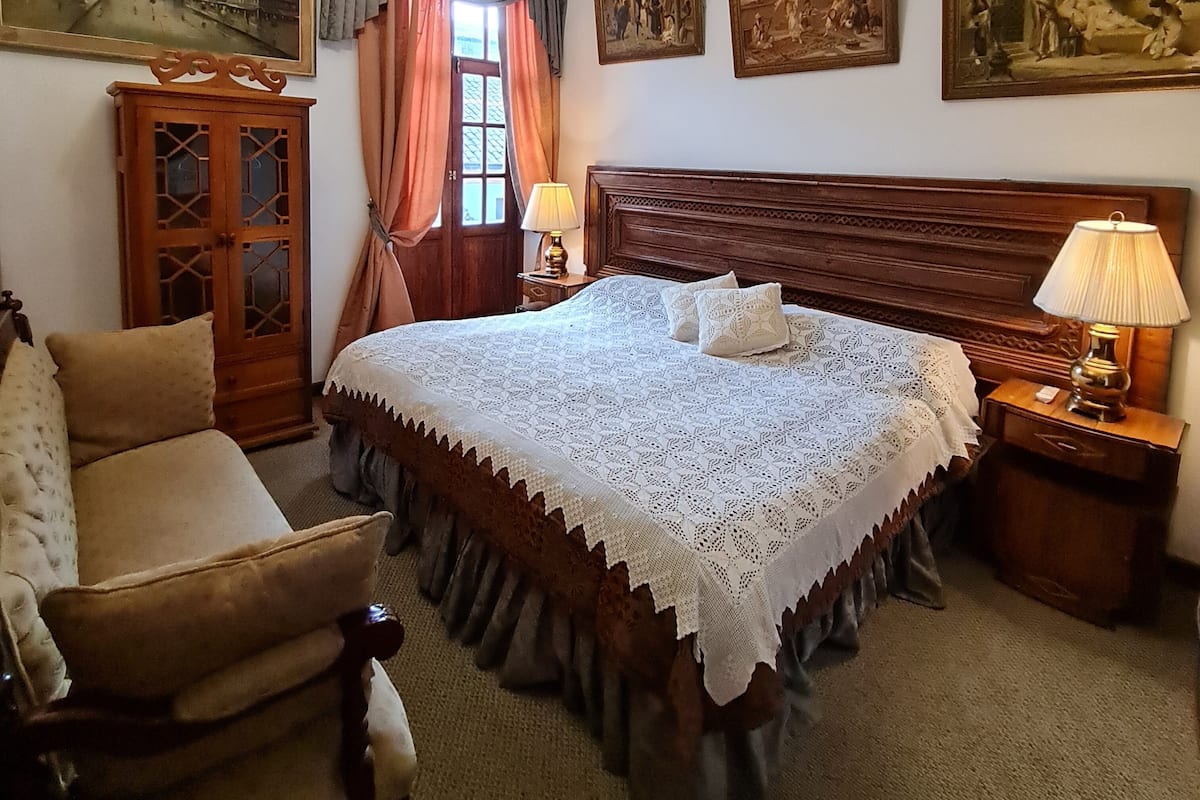A bedroom with a large bed, a wooden headboard, a nightstand with a lamp, a chair, and a wardrobe. There are paintings on the walls and a window with curtains.