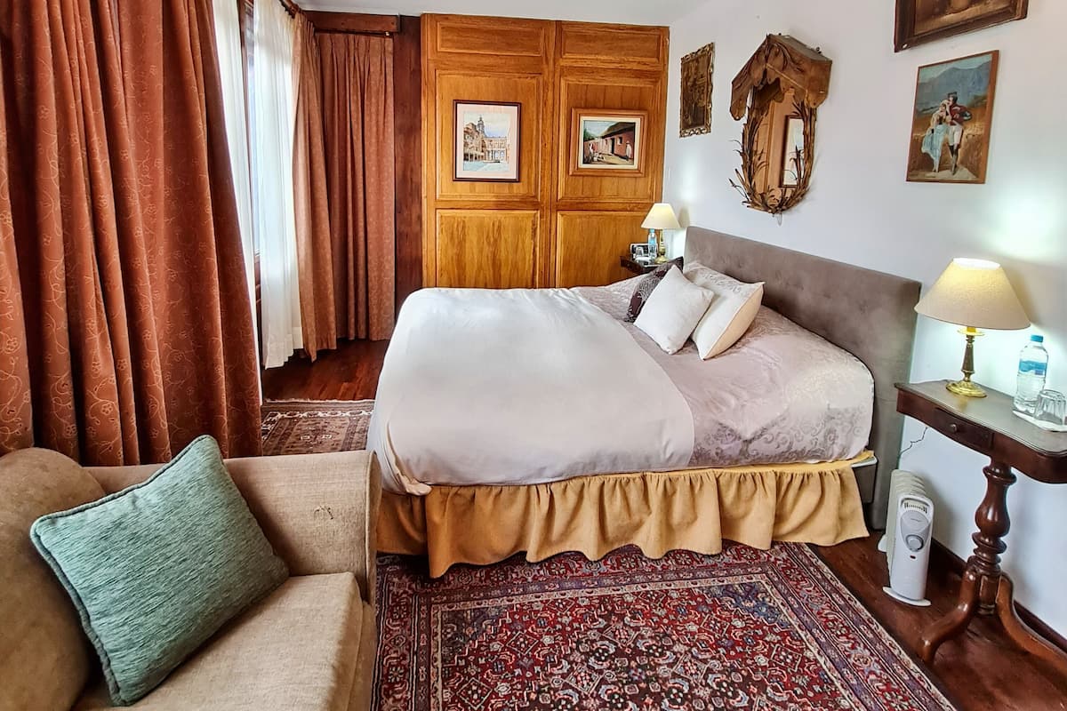 A hotel room with a bed, a bedside table with a lamp, a small table with a bottle and glasses, a decorative mirror, and a patterned rug.
