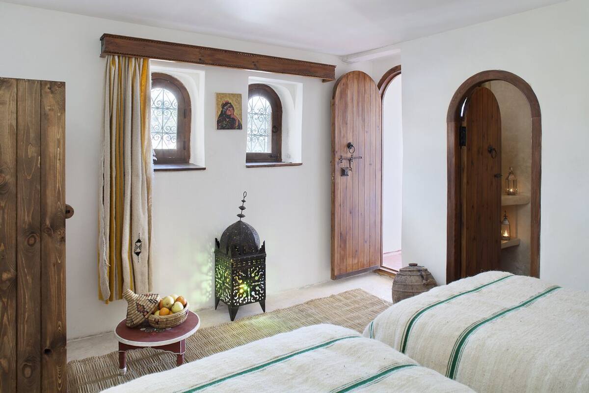 A room with two beds, a wooden door, a window with curtains, a lantern, and a small table with fruit.