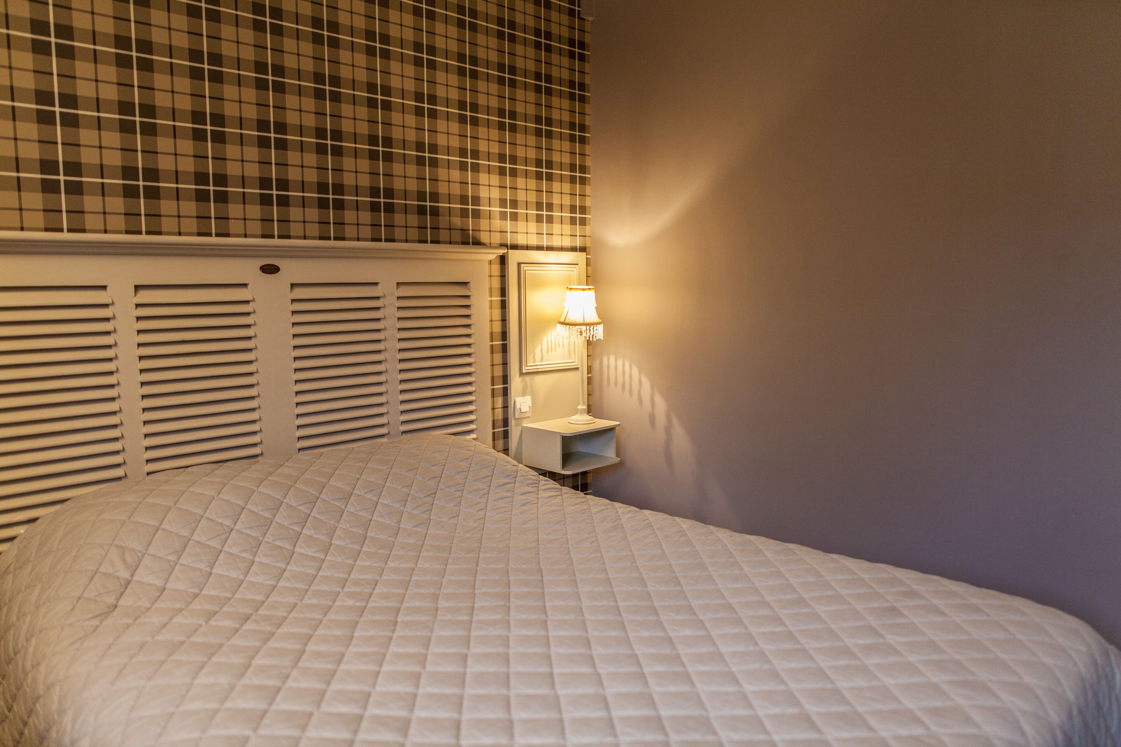 A neatly made bed with a quilted cover, a bedside table with a lamp, and a checkered wall pattern.
