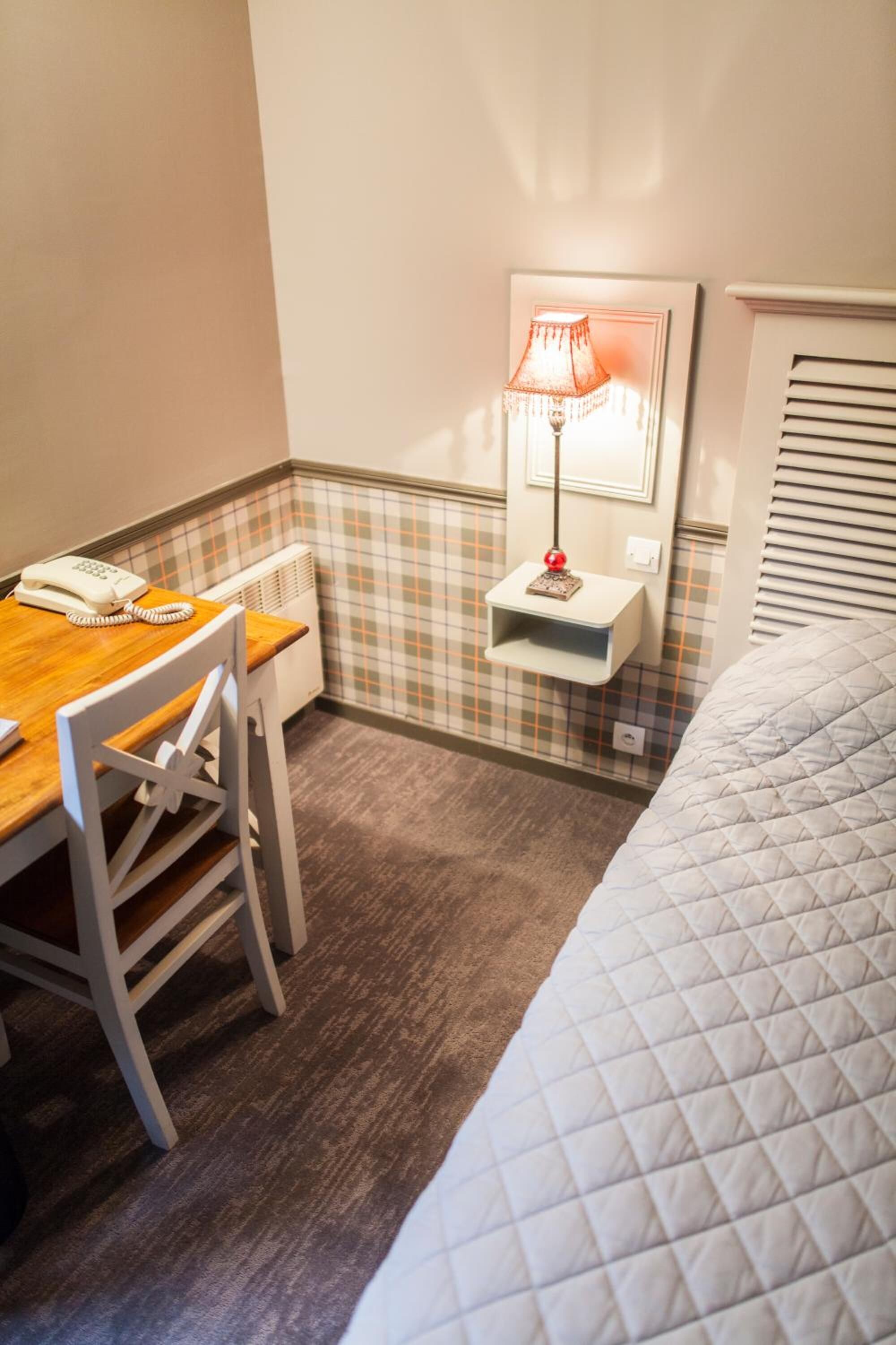 Een hotelkamer met een houten bureau, een stoel, een bed met een gewatteerd dekbedovertrek, een lamp op een plank en een radiator.
