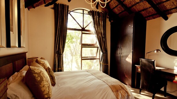 A bedroom with a bed, a desk, a chair, a window with curtains, a chandelier, and a round mirror. - Lourie Lodge (Sandton)