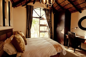 A bedroom with a bed, a desk, a chair, a window with curtains, a chandelier, and a round mirror. - Lourie Lodge (Sandton)