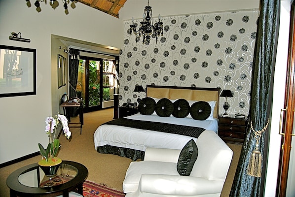 A bedroom with a large bed, a bedside table, a mirror, a chandelier, and a view of the outdoors. - Lourie Lodge (Sandton)