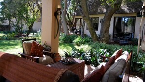 A covered outdoor seating area with wicker furniture, a table with decorative items, and a view of a house with a thatched roof. - Lourie Lodge (Sandton)